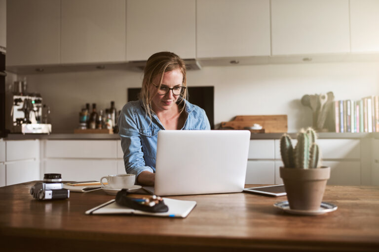 Smiling young female entrepreneur sitting at her kitchen table at home working on her small business with a laptop